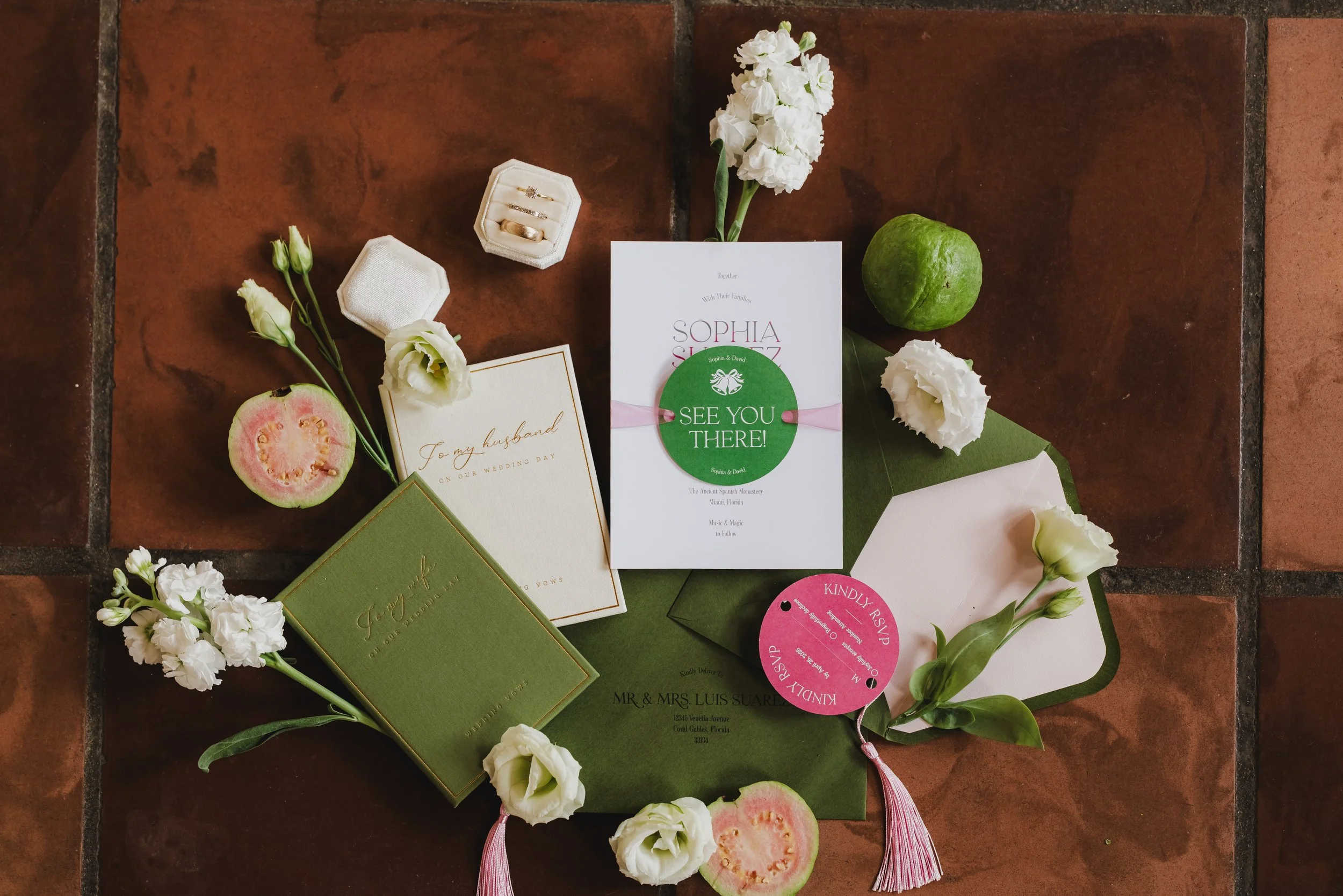 Wedding invitation with white and pink accents, surrounded by white flowers, a green fruit, and slices of guava on a brown tiled surface.
