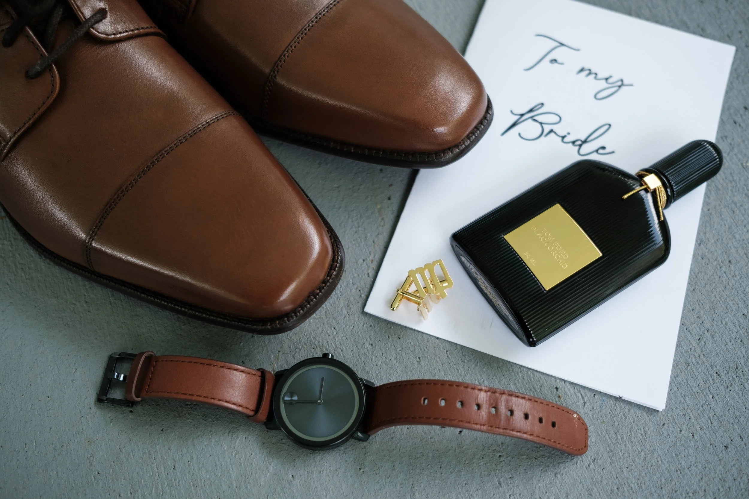 Brown leather dress shoes, a black perfume bottle with gold accents, a black wristwatch with a brown strap, and a white card with handwritten text that says 'To my Bride' on a gray surface.