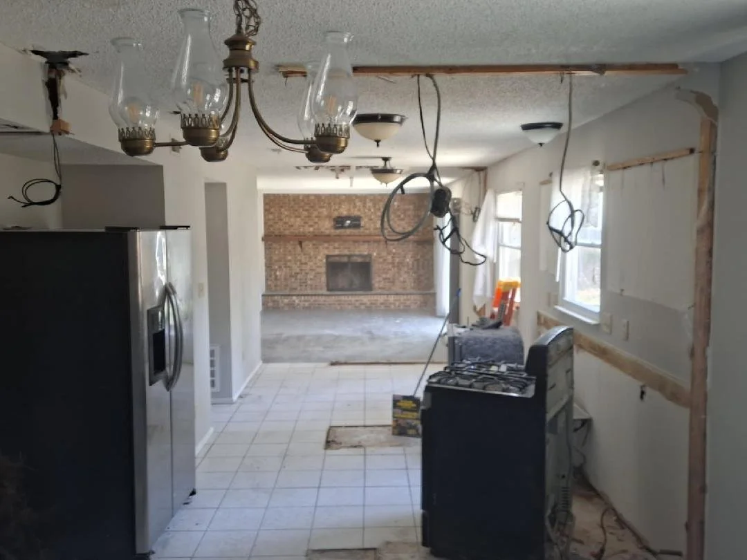 Kitchen undergoing renovation with exposed wiring, missing cabinet doors, and a partially removed wall. A stove is present, and a brick fireplace is visible in the living area.