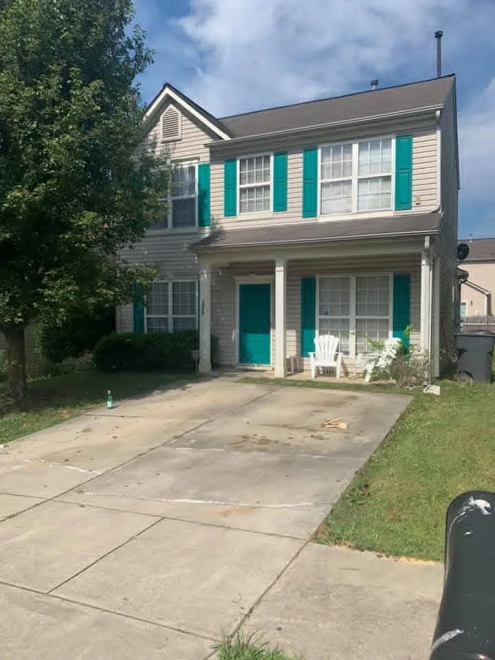 A two-story house with beige siding and teal shutters. The house has a front porch with white chairs and a teal door. There is a driveway and some greenery, including a tree and bushes.