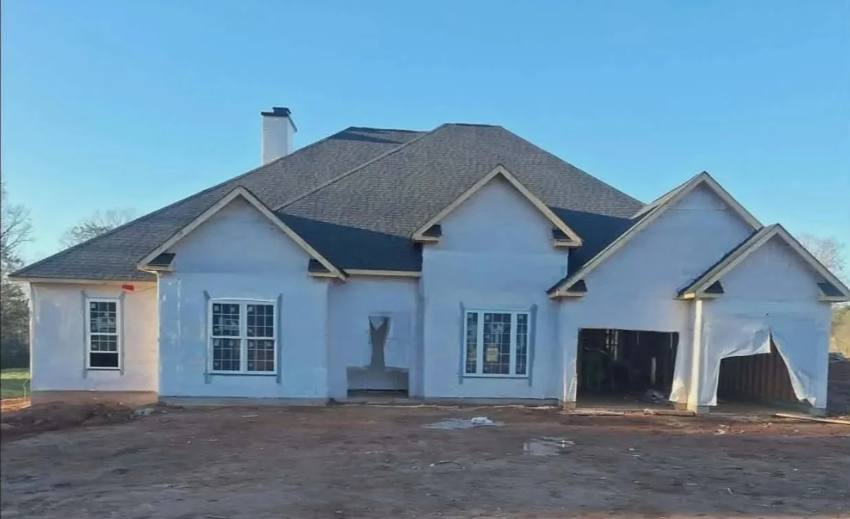 A house under construction with white exterior walls, multiple windows, and an attached garage without doors. The roof is shingled, and the ground around the house is bare dirt.