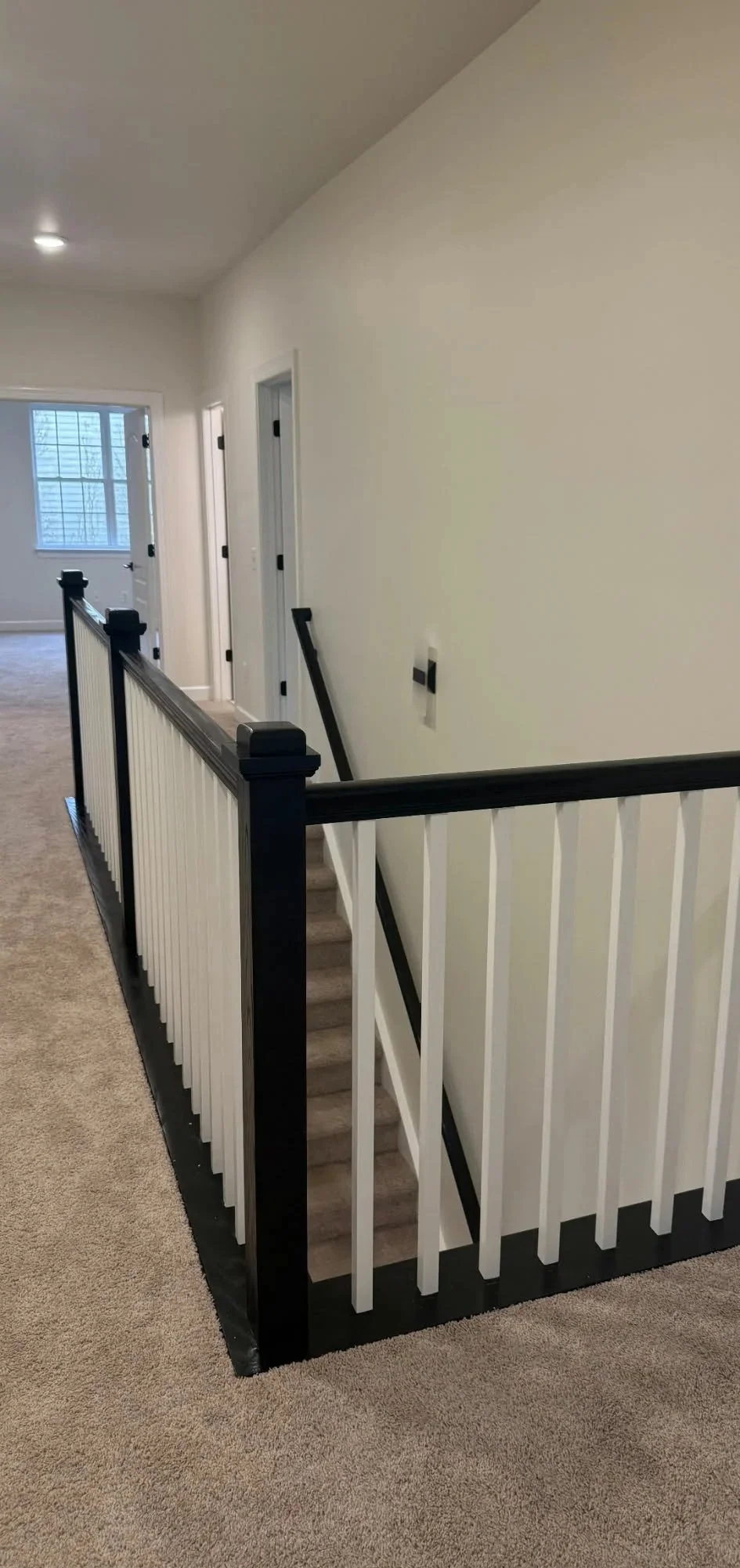 Interior view of a hallway with a staircase, a black and white railing, a beige carpet, and doors leading to rooms. There is a window at the far end and a light on the ceiling.
