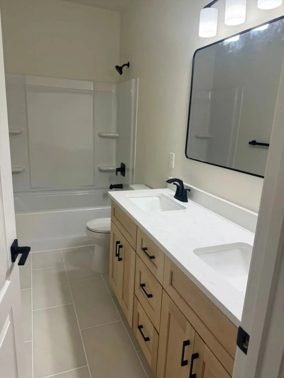 Bathroom with dual sinks, wooden vanity, rectangular mirror, and bathtub with shower, black fixtures, and beige tile flooring.