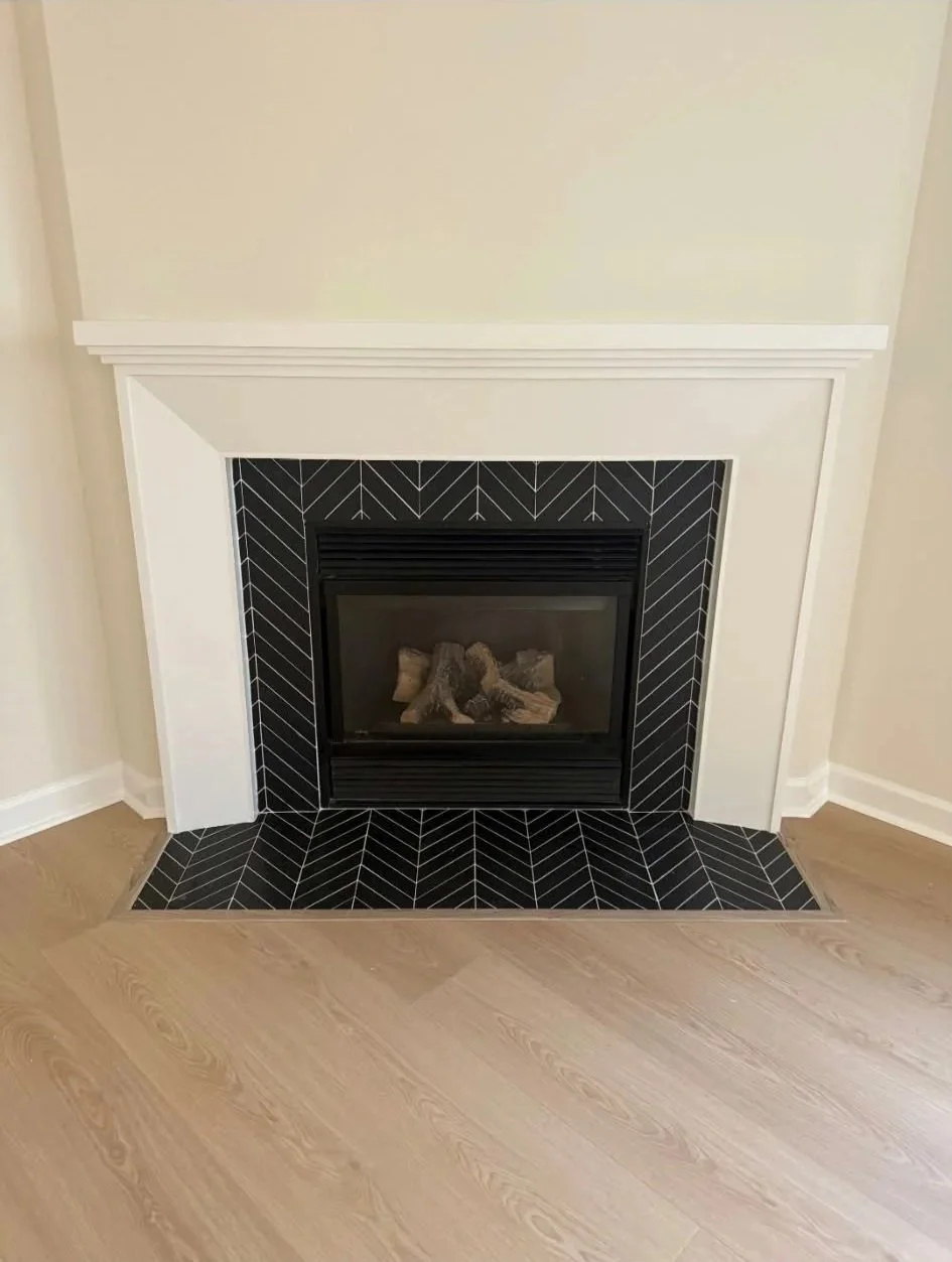 A modern fireplace with a black and white herringbone tile surround and black metal frame, set into a white wall with cream-colored walls on either side and light brown wood flooring.
