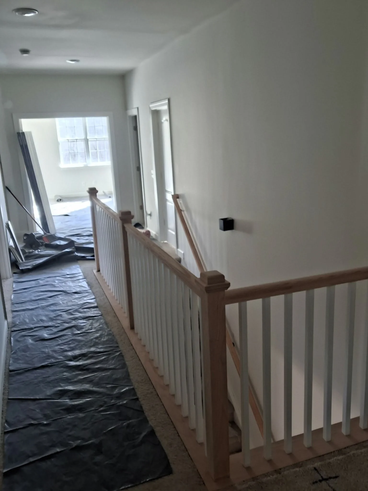 Interior view of a house under construction or renovation, showing a staircase with light wood and white balusters, black plastic floor covering, and construction tools and materials near an open stairway and closed doors.
