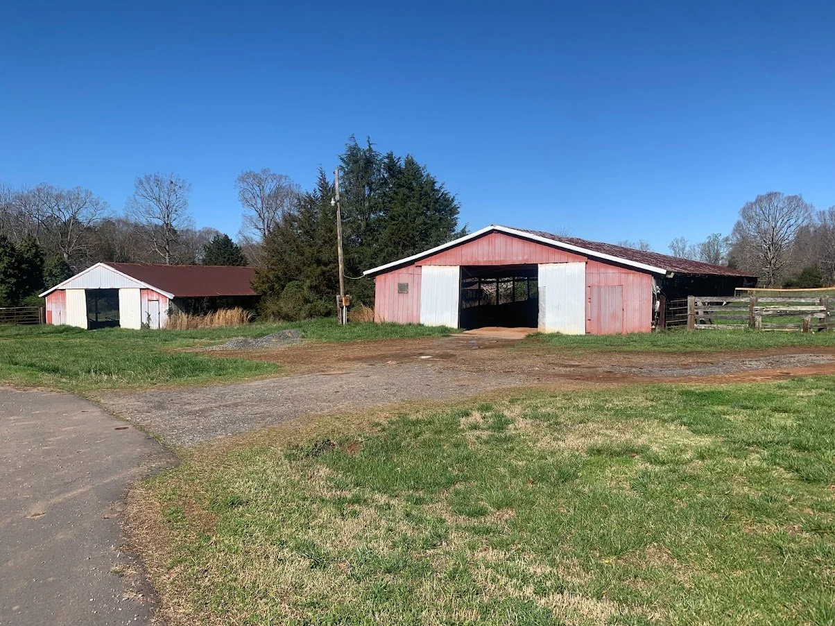 Two red and white farm buildings on a field with green grass and trees in the background, clear blue sky.
