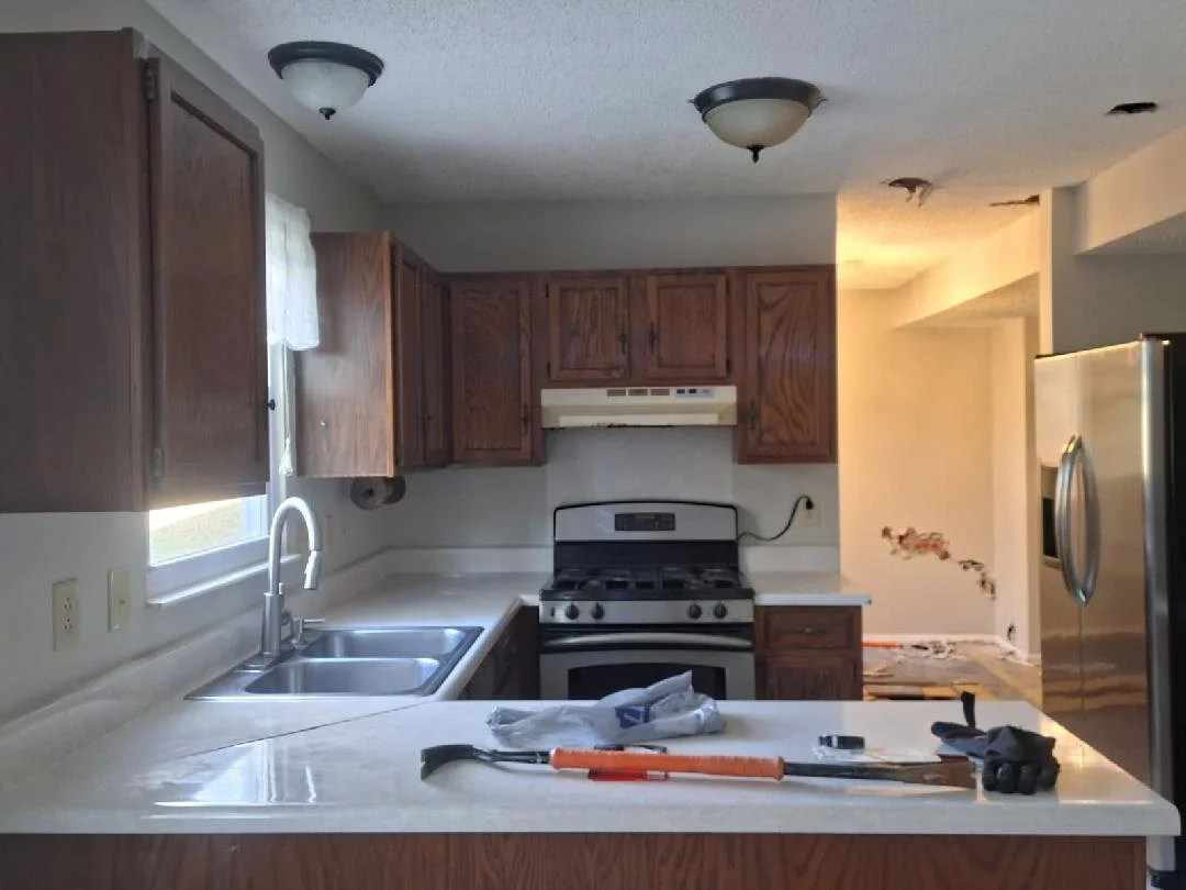 Kitchen with wooden cabinets, white countertops, a window with a curtain, stainless steel sink, stove, and refrigerator, with tools and items on the counter.
