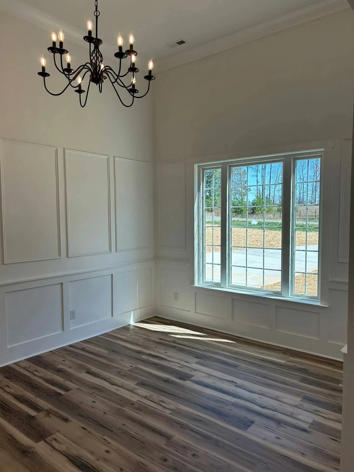 Empty room with white paneled walls, a large window, wood flooring, and a black chandelier.