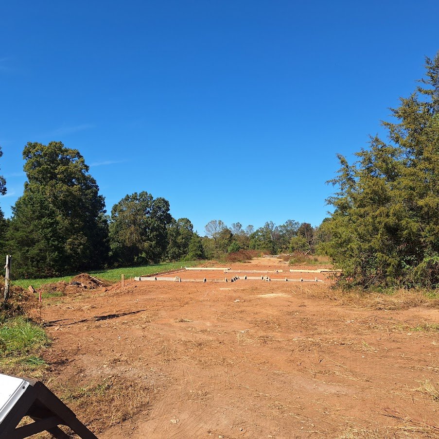 A construction site with cleared land and some foundation or footing structures being built, surrounded by trees under a bright blue sky.