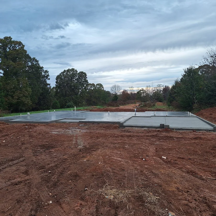 Newly poured concrete foundation on a construction site surrounded by dirt and trees under a cloudy sky.