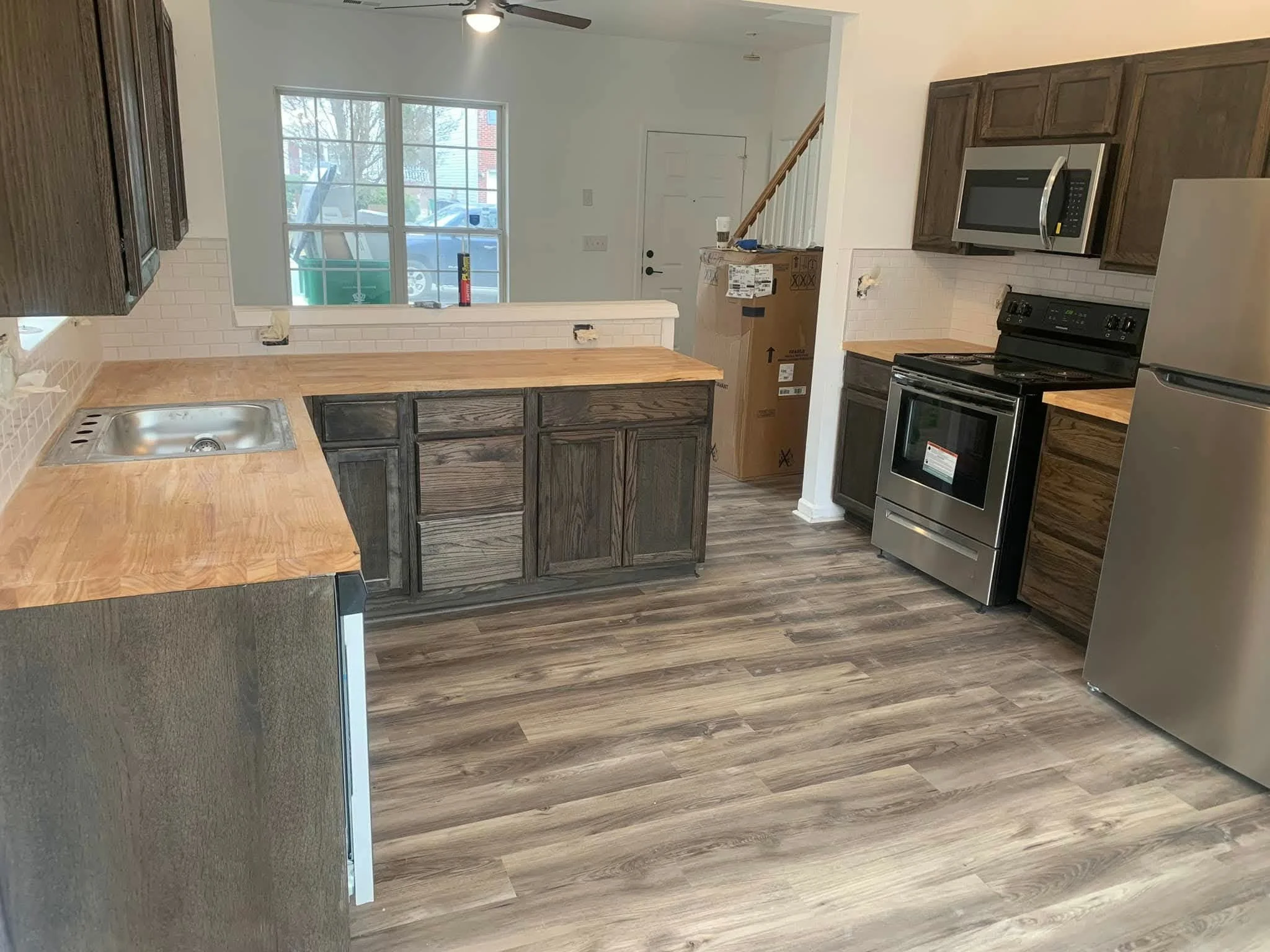 Empty kitchen with wooden floors, dark wood cabinets, stainless steel refrigerator, oven, microwave, and beige countertops, with a window overlooking the outside.