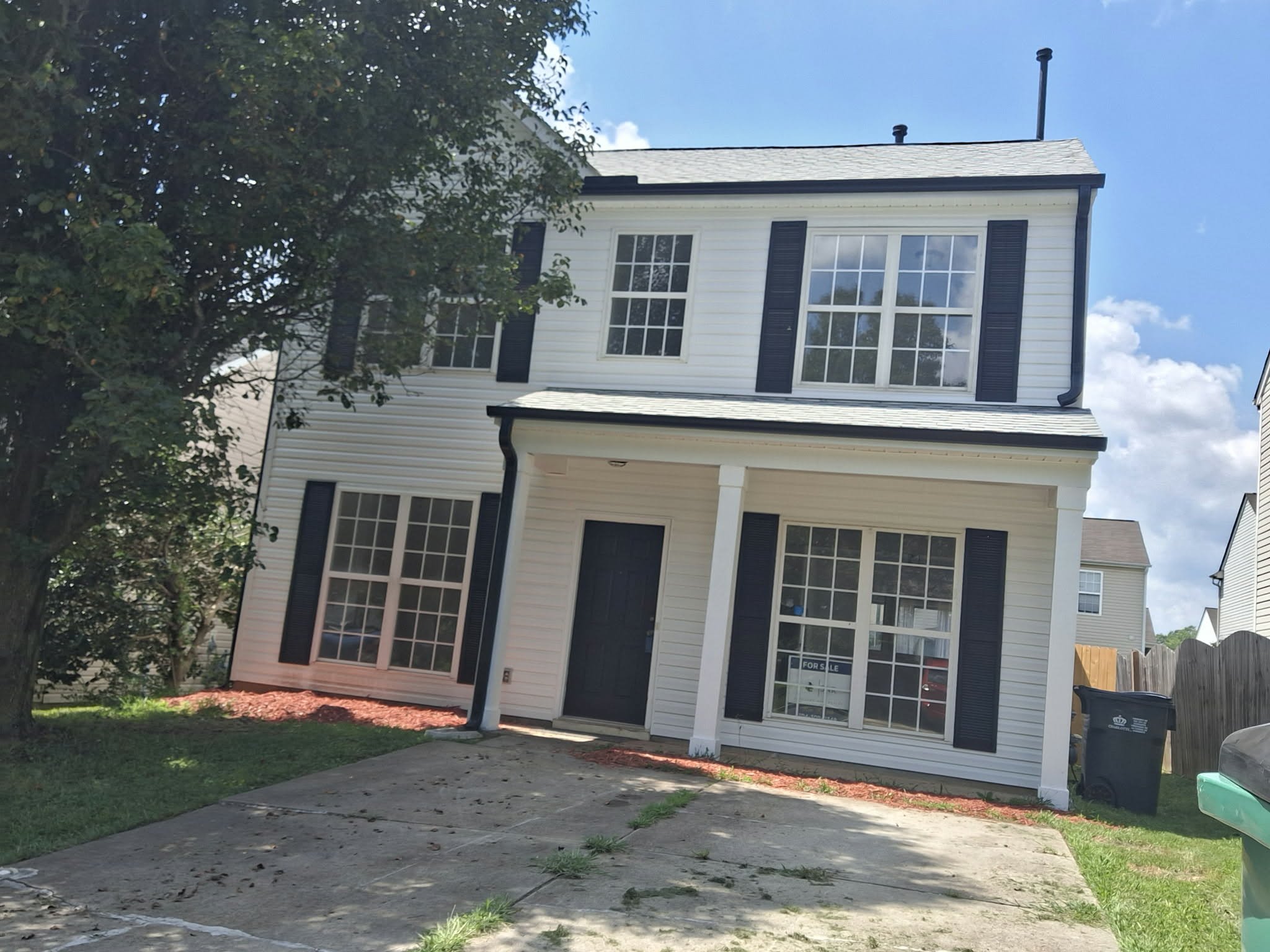 A two-story white house with black window shutters, a black front door, and a concrete driveway. There is a large tree on the left side and a wooden fence on the right. A "For Sale" sign is visible in the front window on the right.
