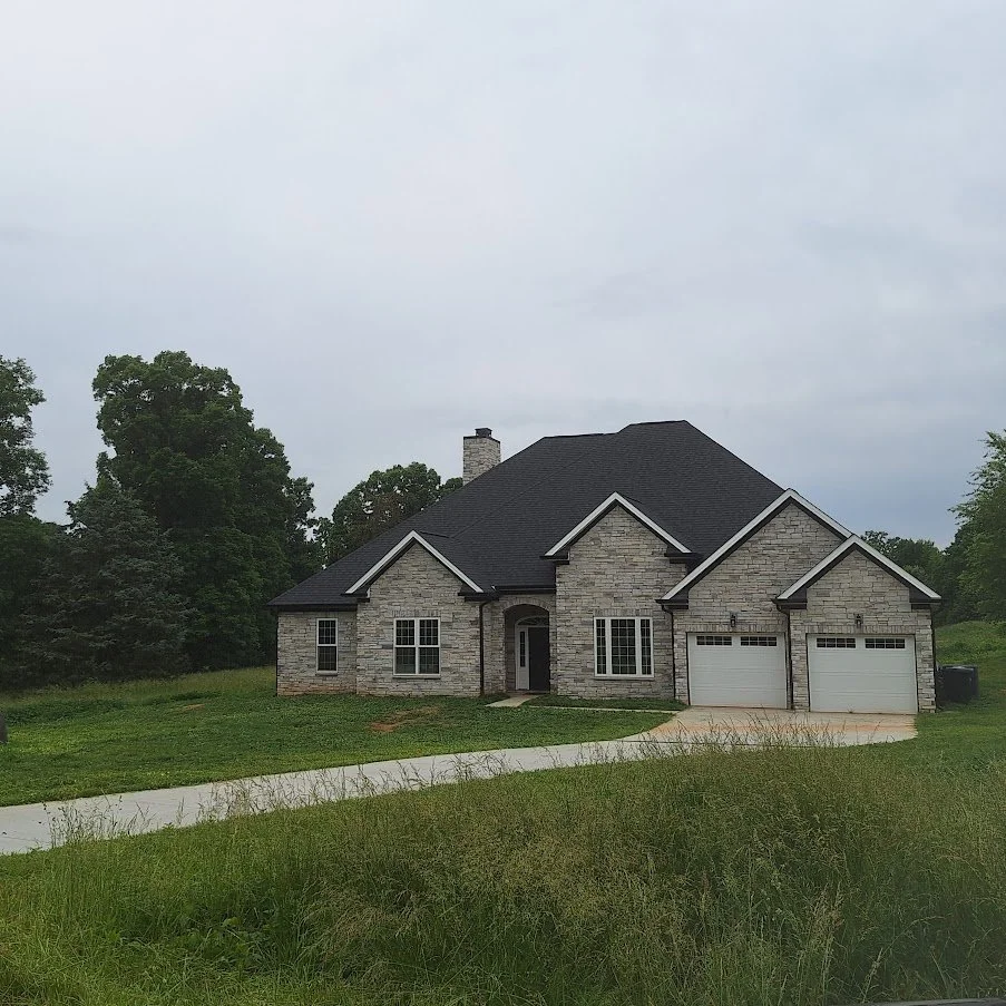 A two-story house with a stone exterior, black roof, front lawn, and a driveway with two garage doors.
