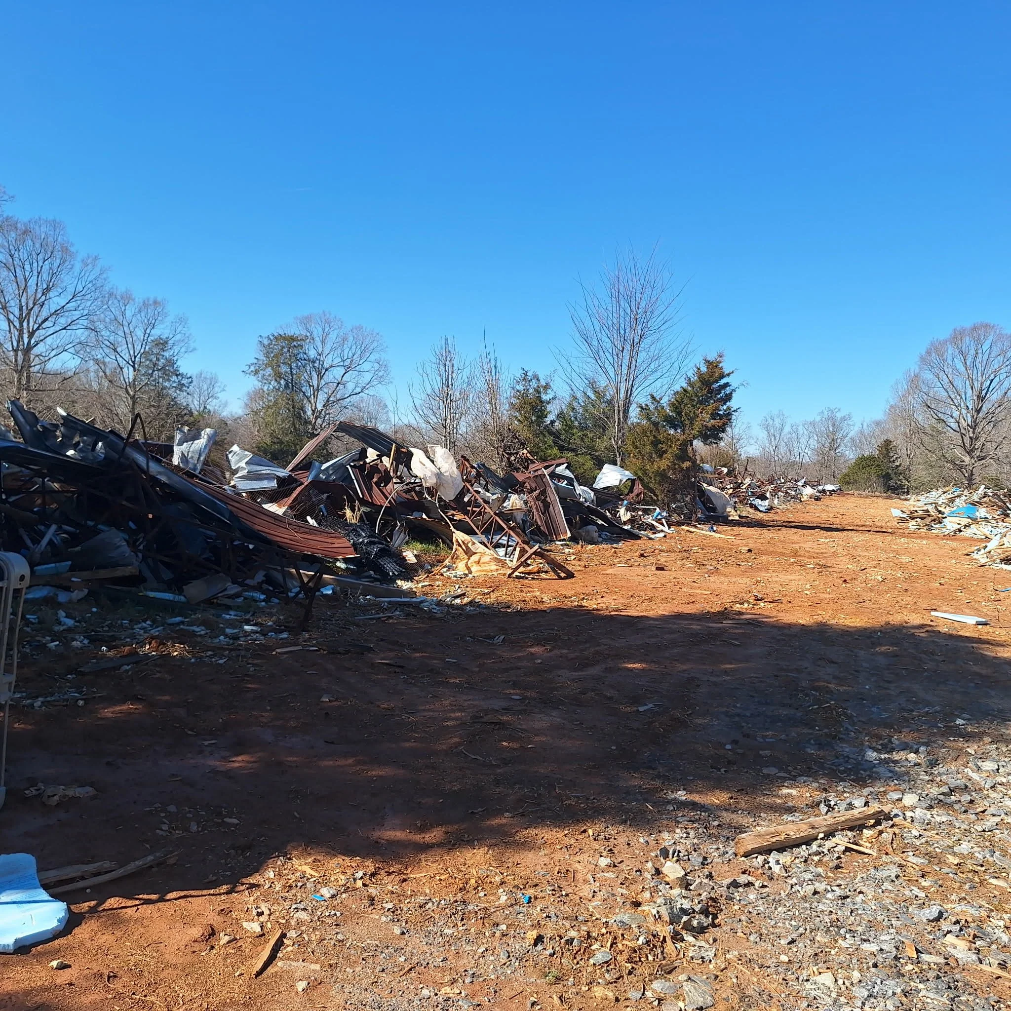 Debris and wreckage of destroyed structures spread across a dirt field with leafless trees in the background under a clear blue sky.