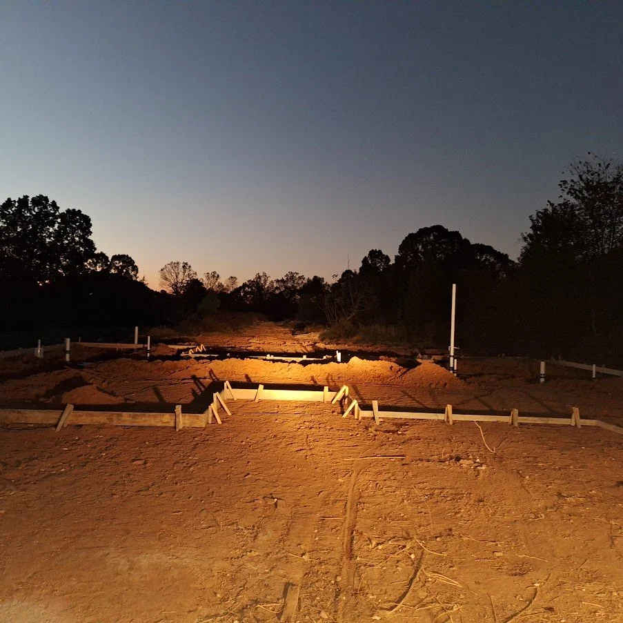 Construction site at dusk with wooden frames and markers on a dirt path, surrounded by trees and a clear sky
