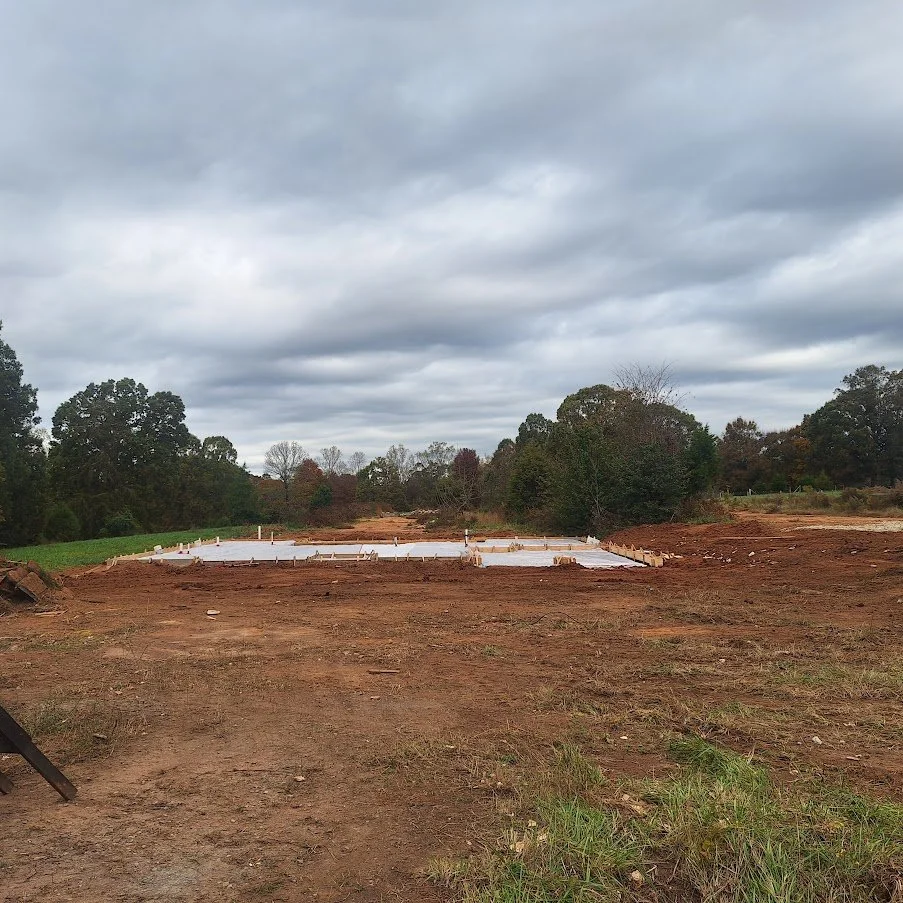 Construction site with a foundation covered in white plastic, surrounded by cleared dirt and trees under a cloudy sky.