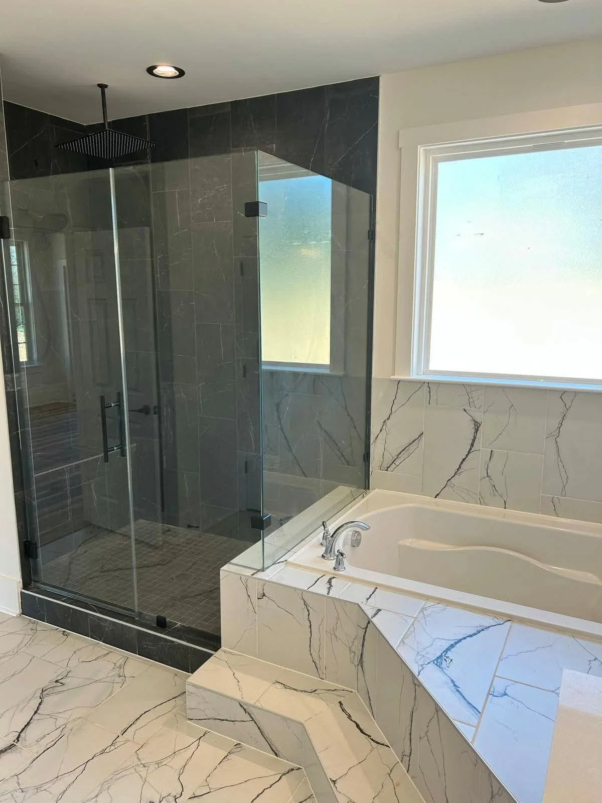 Modern bathroom with a glass-enclosed shower, a large window, and a built-in bathtub with marble-like tiling.