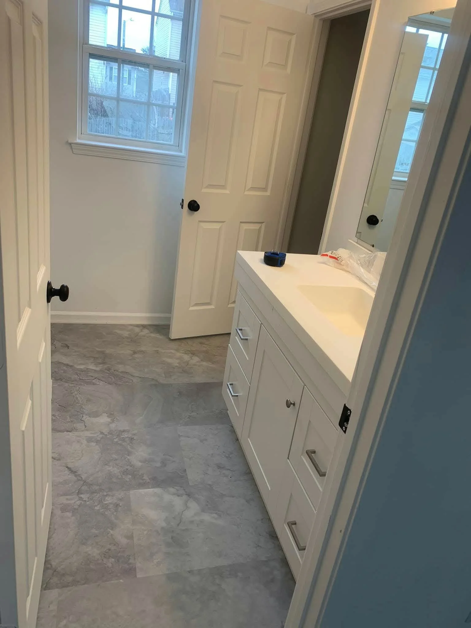 Small bathroom with white vanity, mirror, and marble floor tiles. Window shows neighboring houses.