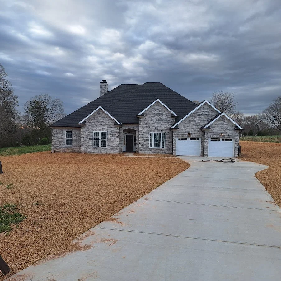 Newly built gray brick house with two-car garage and dark shingled roof on a cloudy day, with a curved concrete driveway and a bare front yard.