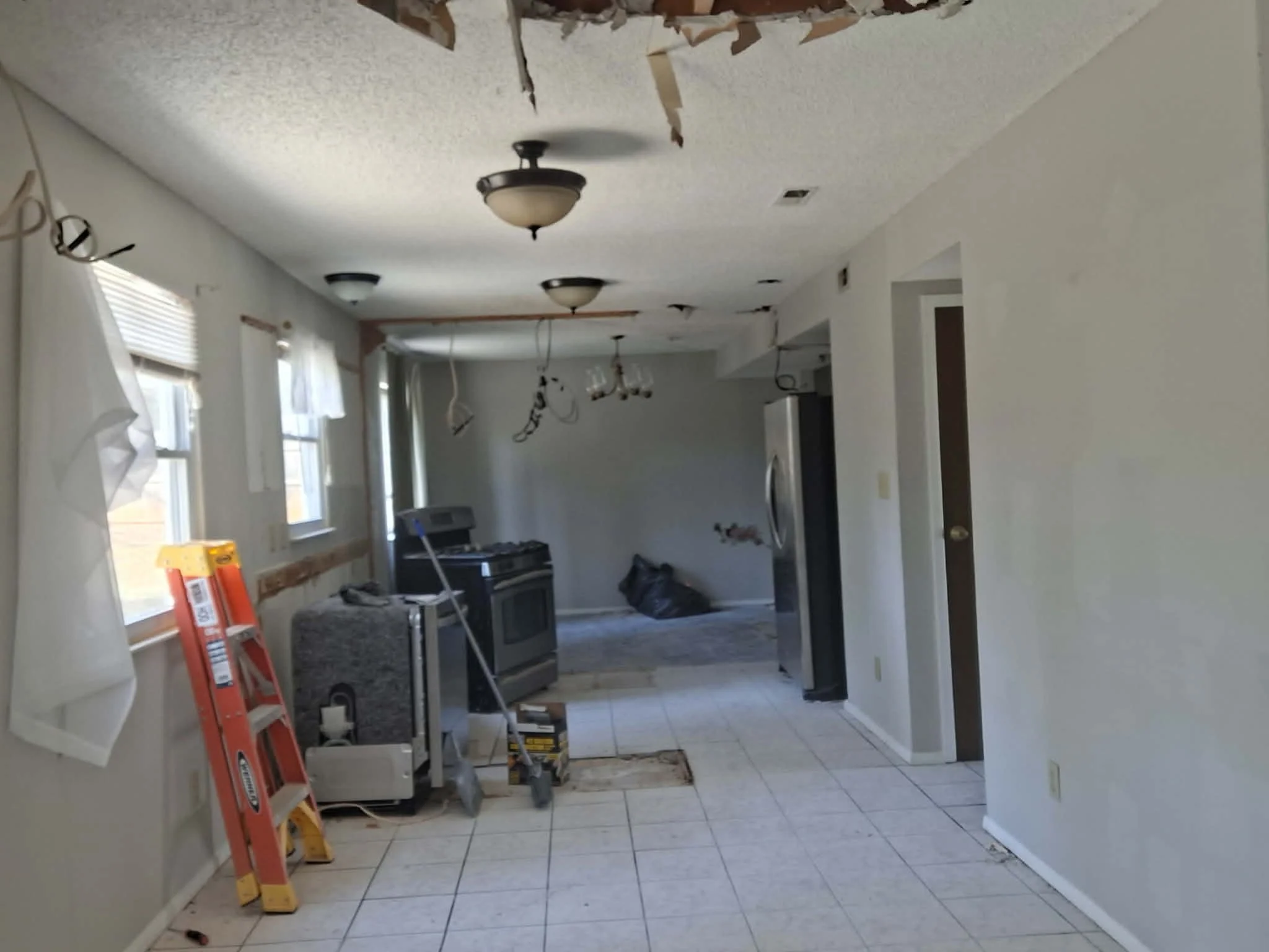 A kitchen undergoing renovation with torn ceiling, exposed wiring, and construction tools. There's a ladder, mop, and an appliance near the window, and the ceiling light fixtures are missing or damaged.