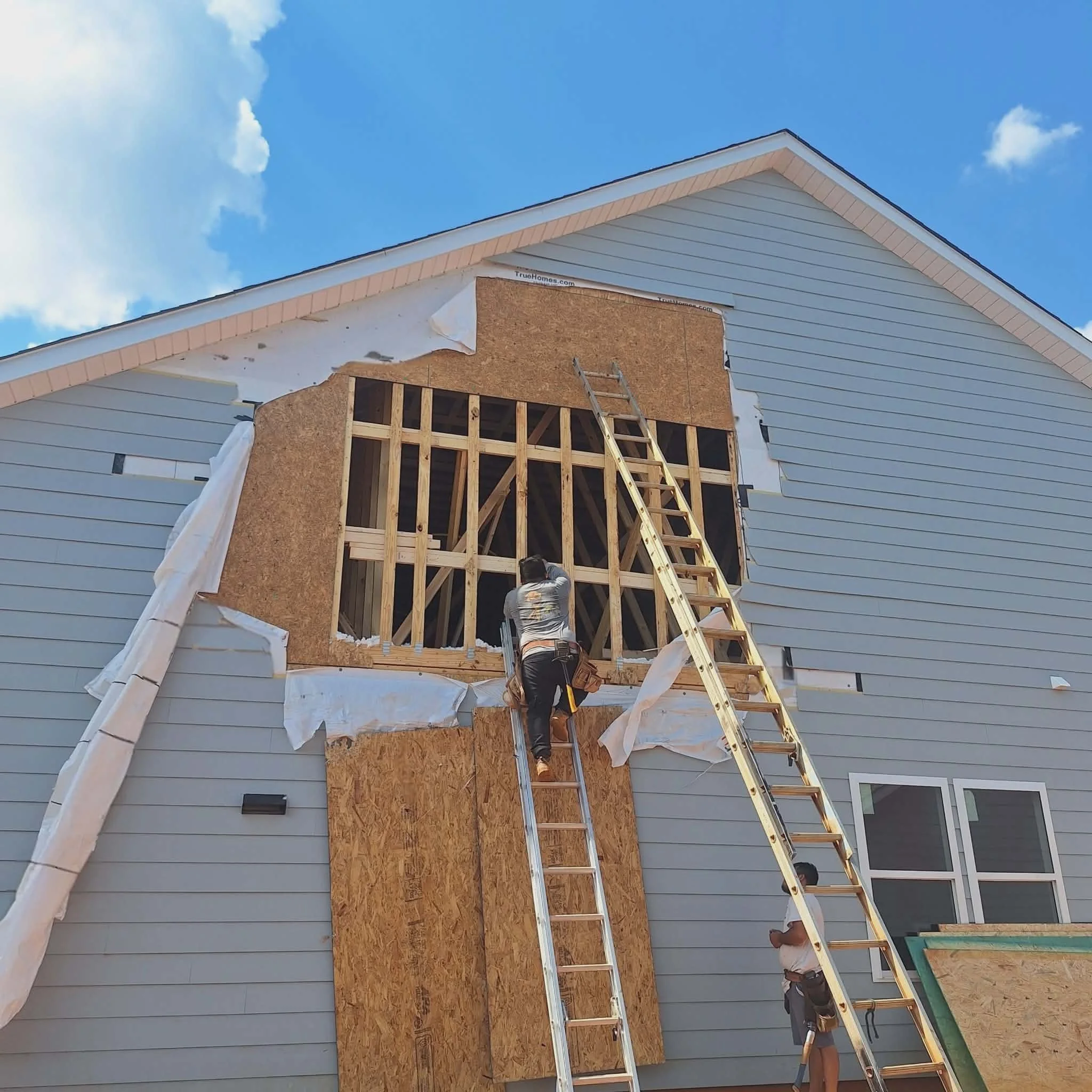 Construction workers installing a large window on the second story of a house with light blue siding, using ladders, with a partly cloudy sky in the background.