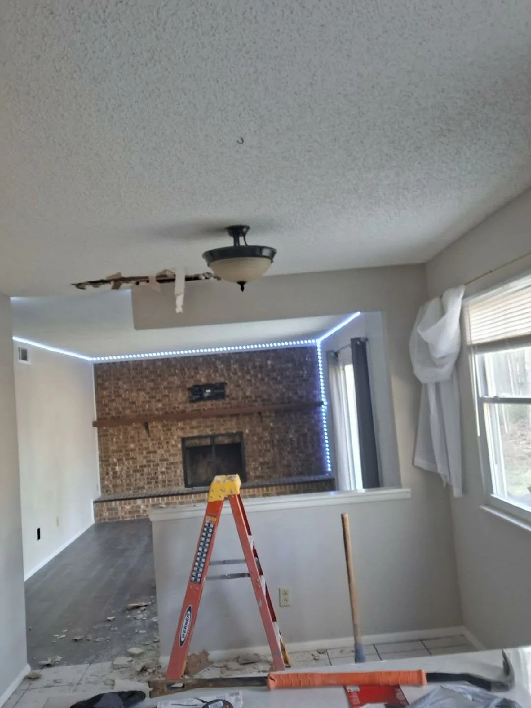 Living room under renovation with a ceiling fan, partially damaged ceiling, brick fireplace, windows with white curtains, and construction tools.