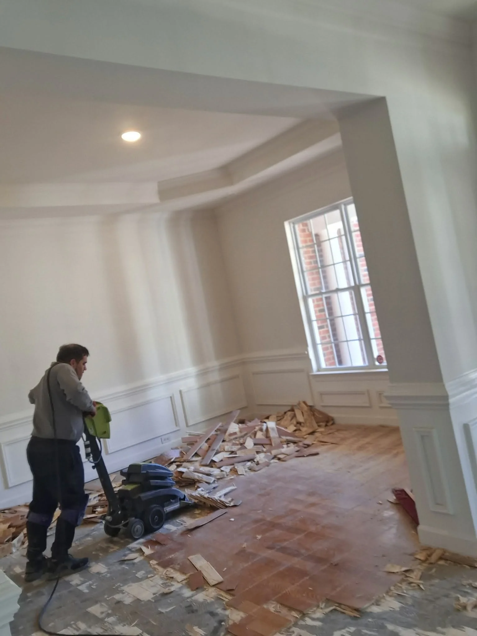 A man using a floor sander on a hardwood floor in a room under renovation, with piles of debris and reclaimed wood on the floor.