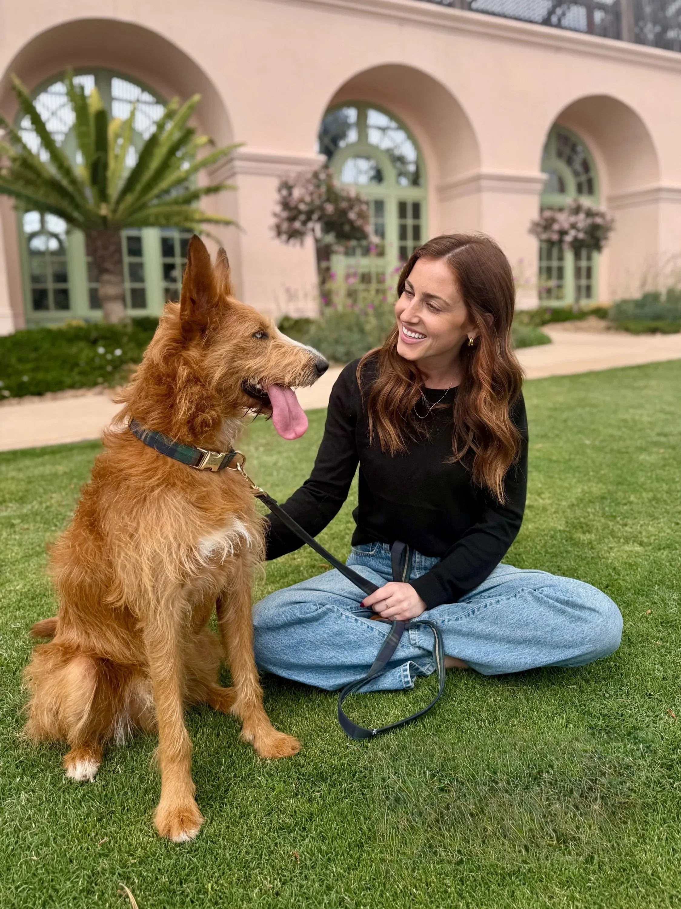 A woman with Auburn hair sitting cross-legged on the grass, smiling at a dog with his tongue sticking out, in front of a beige building with arched windows and flower pots.