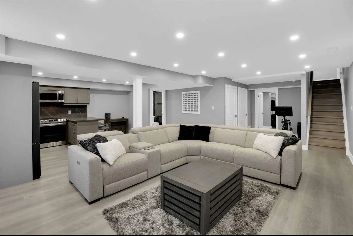 Living room with beige sectional sofa, black and white pillows, a gray coffee table, and a gray carpet. Open kitchen with stainless steel appliances, gray cabinets, and a small table. Stairs leading up on the right side, with a piece of equipment near the wall.