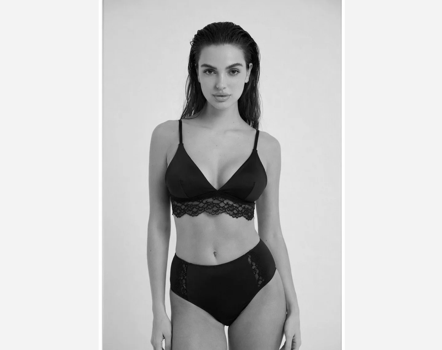 Black and white photo of a woman in lingerie, wearing a lace-trimmed bralette and high-waisted briefs, standing against a plain background.