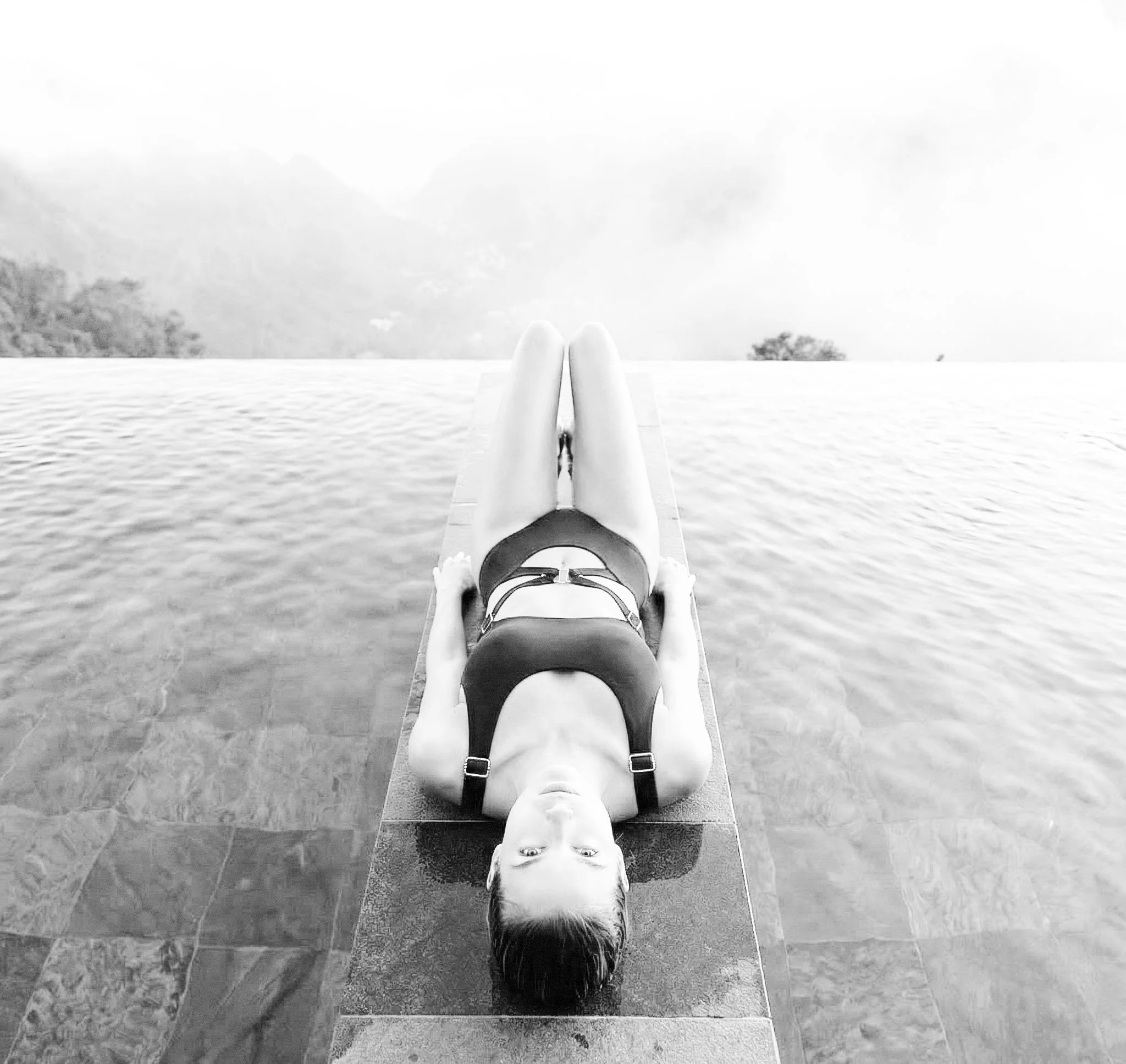 A person lying on the floor outdoors with their head upside down, legs extended, and feet crossed, next to a body of water and mountainous landscape in the background.