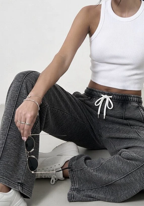 Person sitting cross-legged on the floor, wearing a white crop top, dark gray sweatpants, white sneakers, and holding sunglasses.