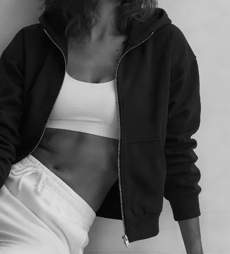 Original designs by athleisure designer Alina. Close-up of a woman wearing a black zip-up hoodie, white crop top, and white sweatpants, showing her midsection.