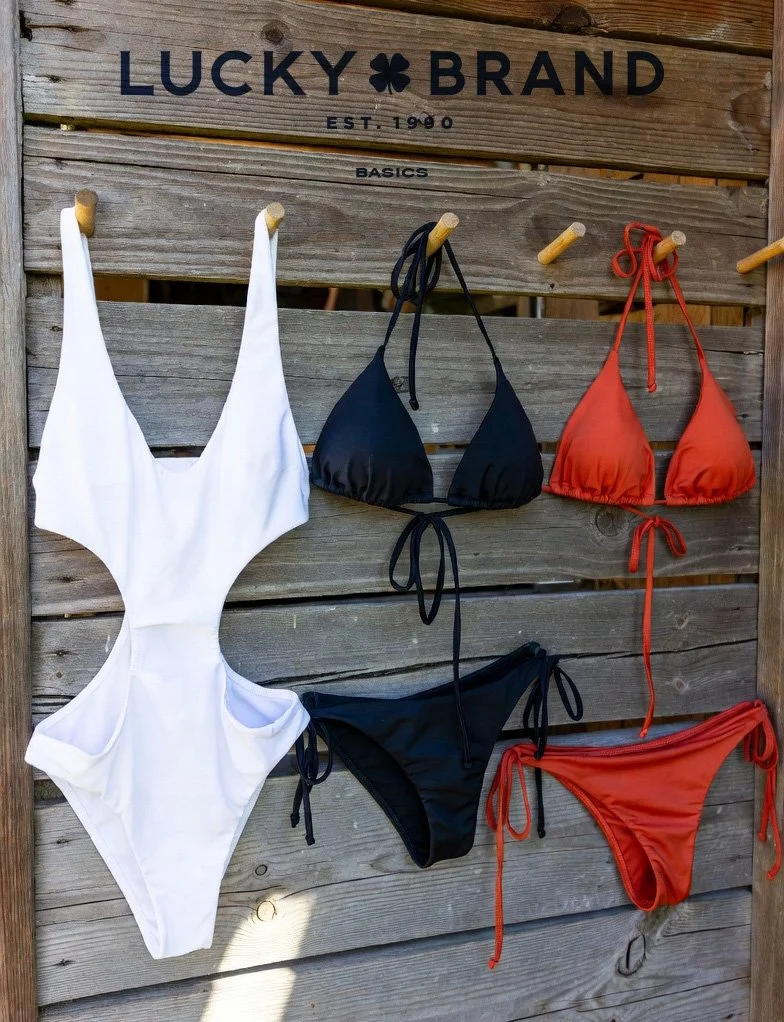 Display of three bikinis hanging on a wooden board, with a white, black, and red swimsuit from the Lucky Brand store, established in 1990.
