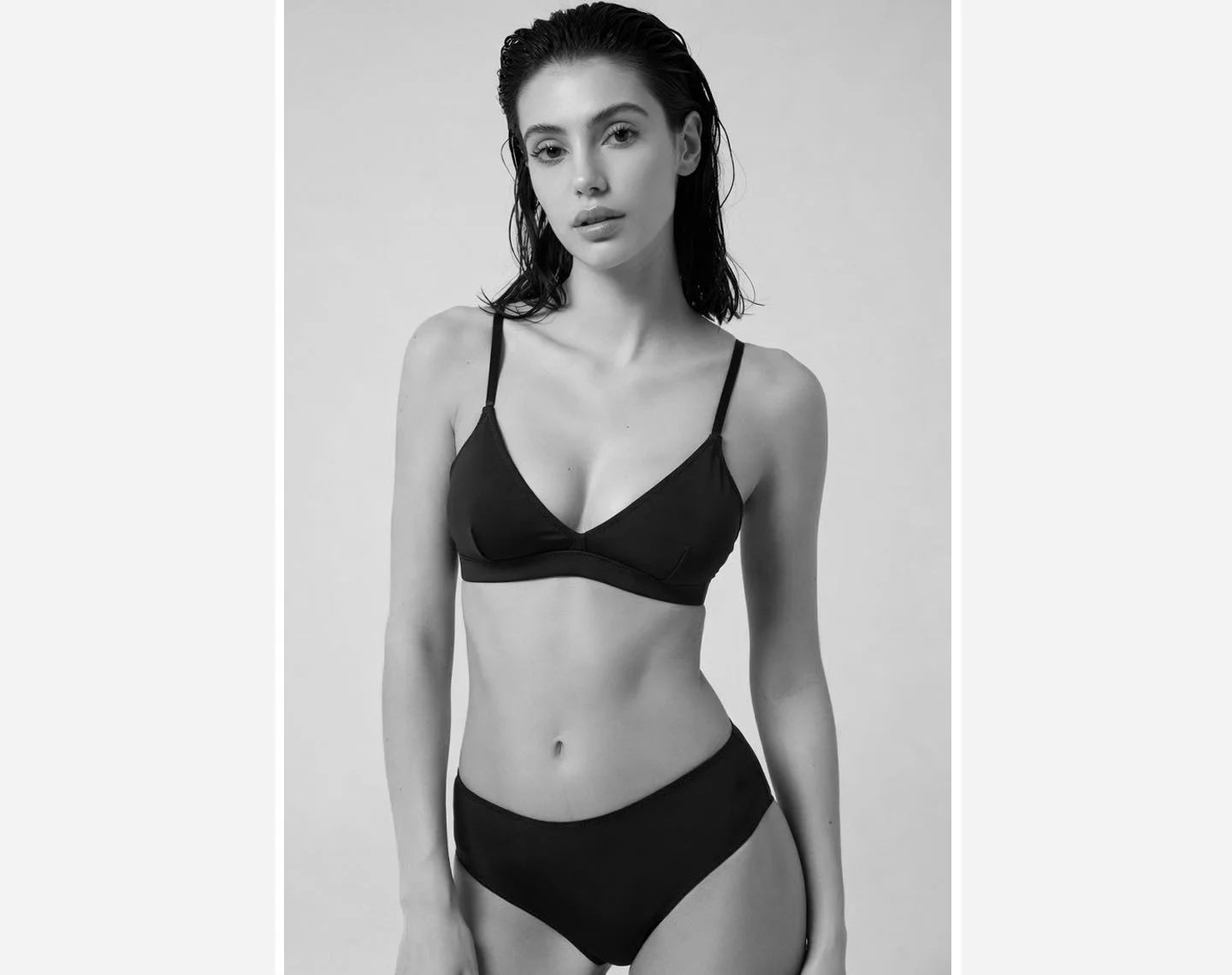 A black-and-white photo of a woman with wet hair, wearing black lingerie, standing against a plain background.