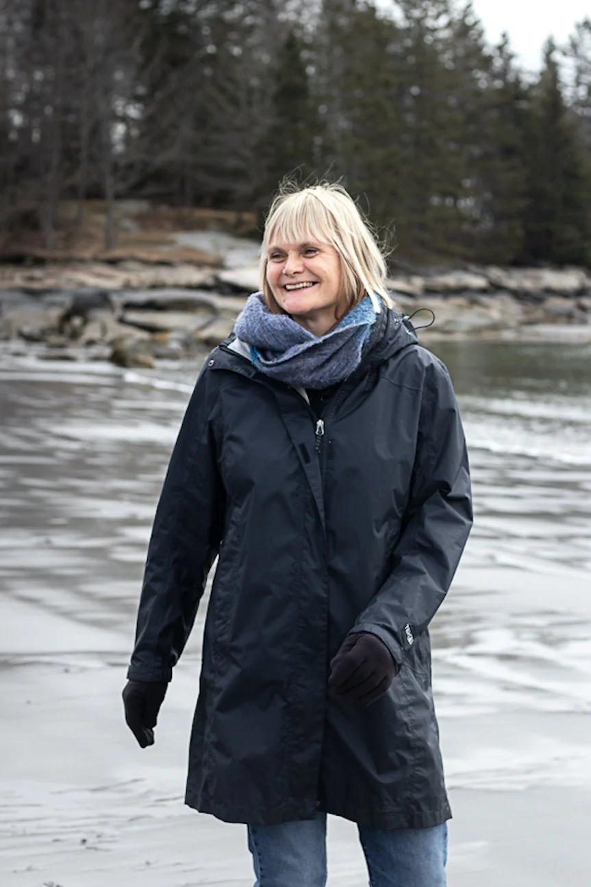 Erika Manning smiling in a black raincoat standing near a body of water with trees in the background.