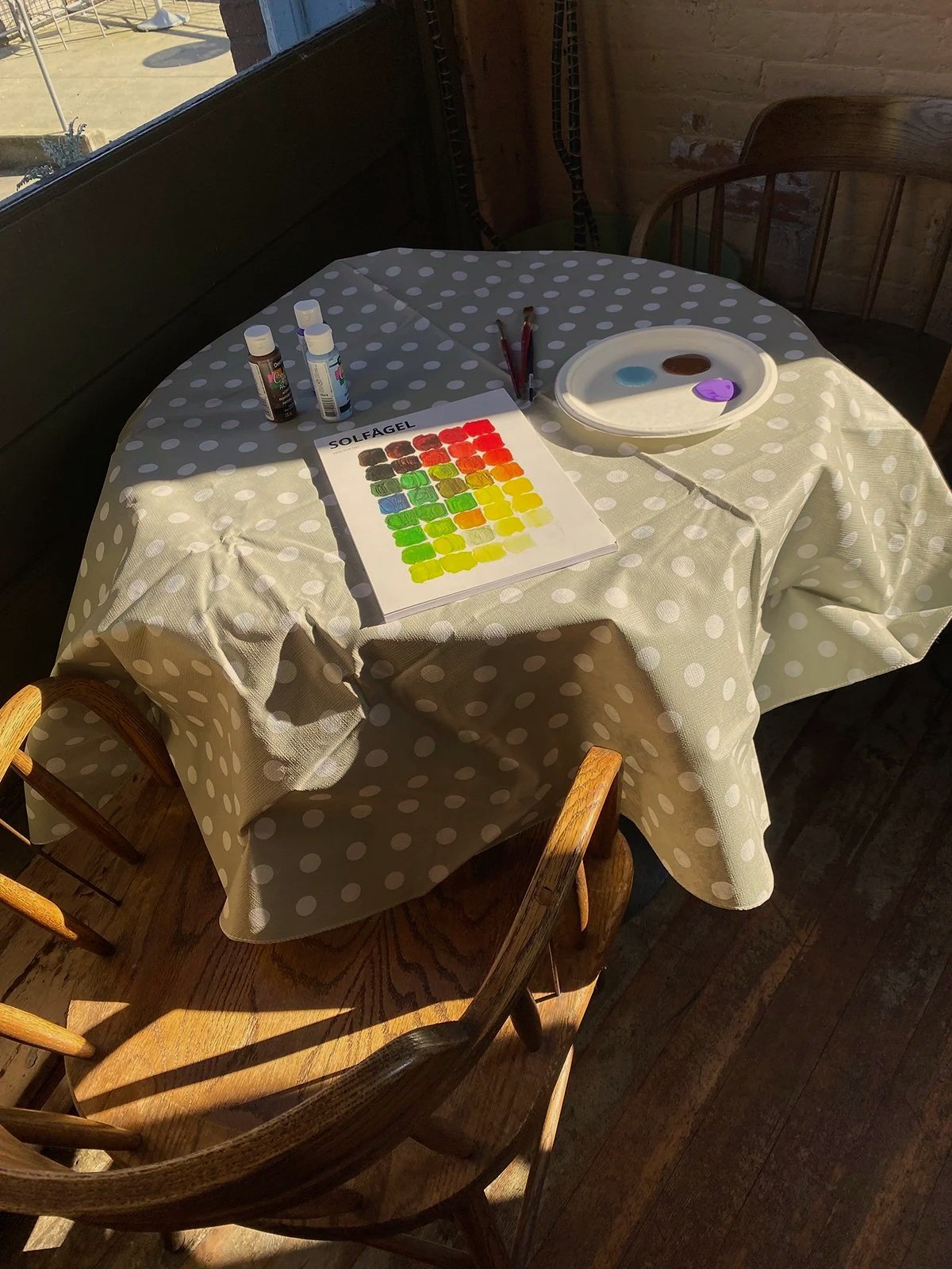 A round table with a grey and white polka dot tablecloth set for paint mixing, featuring bottles of paint, a palette with three colors, and a color mix chart titled 'SOLFAGEL' under a window with sunlight, with wooden chairs around the table.
