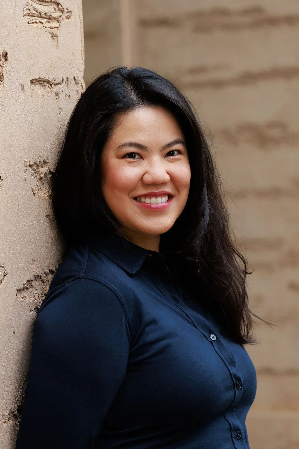 A smiling woman with long dark hair, wearing a navy blue shirt, leaning against a textured beige wall.