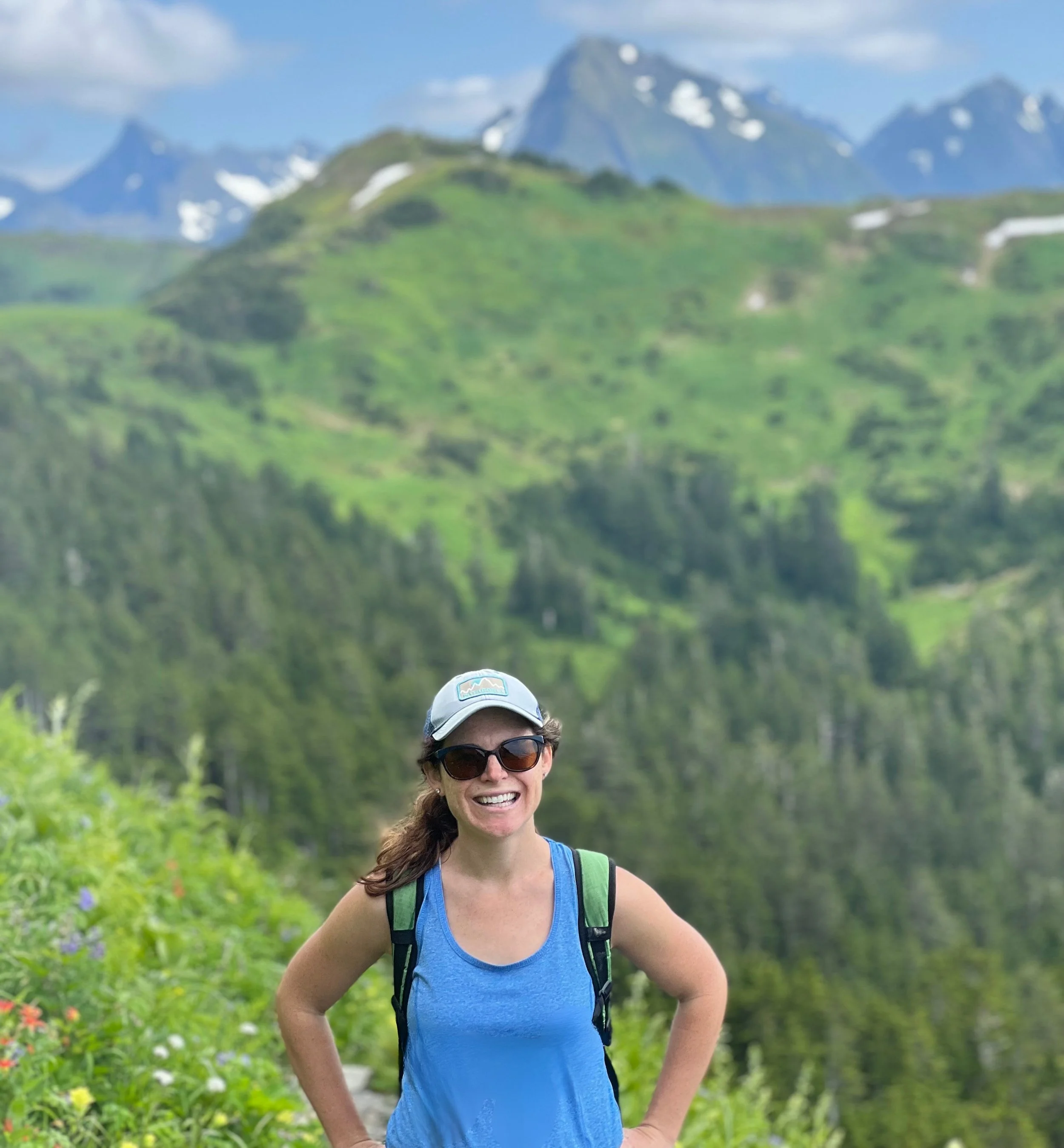 Tiffany Callaway, LCSW, wearing a backpack and smiling outdoors with lush green Alaskan mountains in the background.