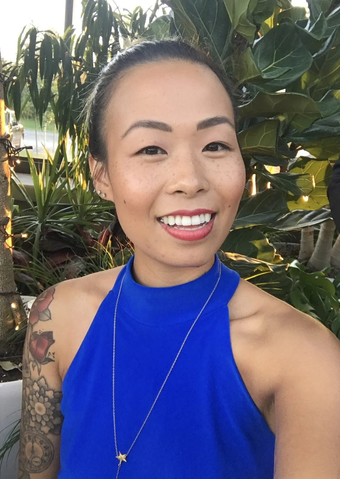 A woman smiling outdoors with lush green plants in the background, wearing a bright blue sleeveless top and a star-shaped necklace.