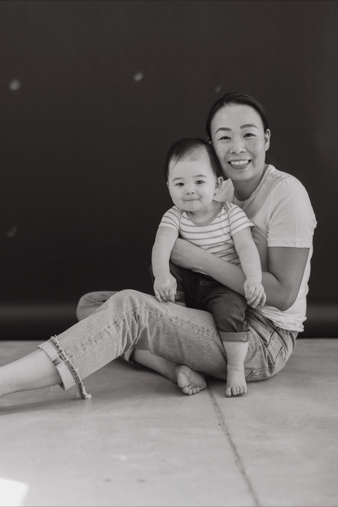 A black and white photo of a smiling woman and a young child sitting on the floor, with the woman holding the child on her lap against a plain dark background.