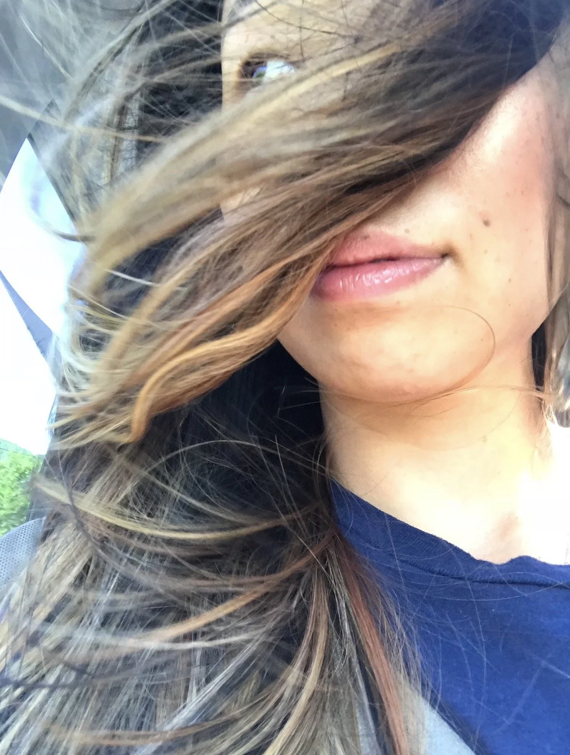 Close-up of a woman's face partially covered by her wavy, light brown hair, showing her lips, part of her nose, and a small mole on her cheek.