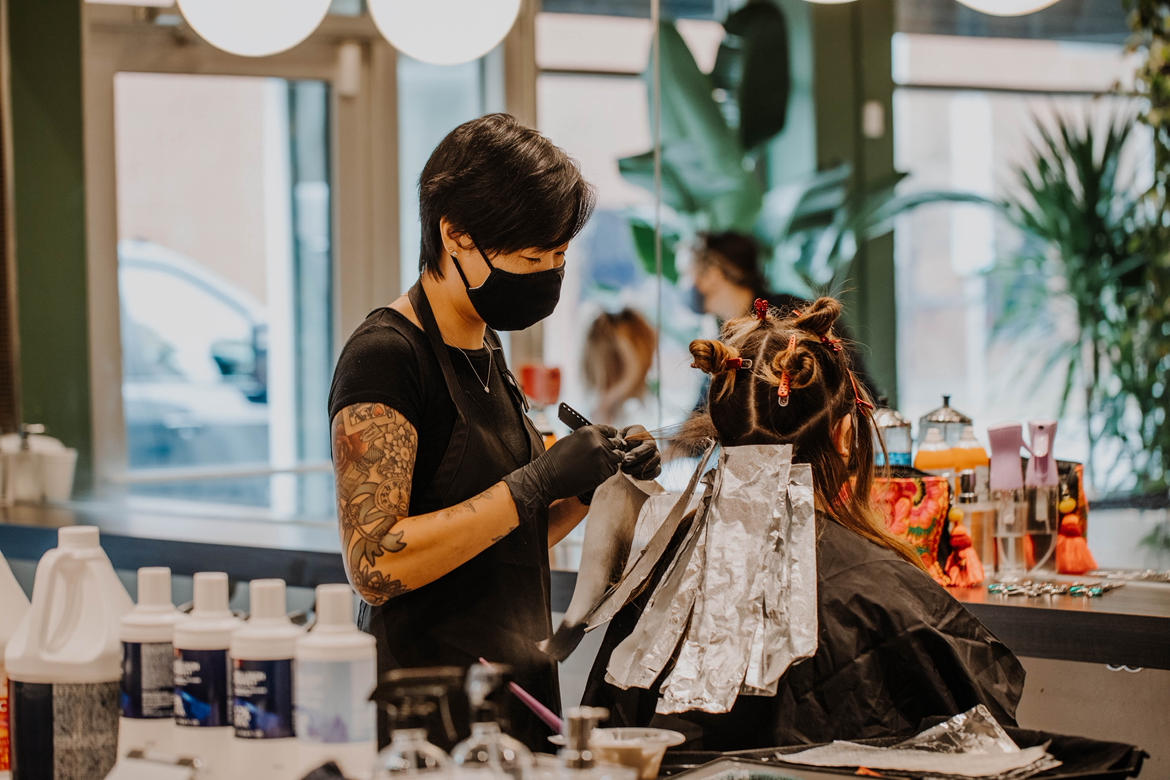 A hairstylist with tattoos on her arms and wearing a black mask and gloves, working on a client's hair in a salon, with various hair products and tools on the counter.