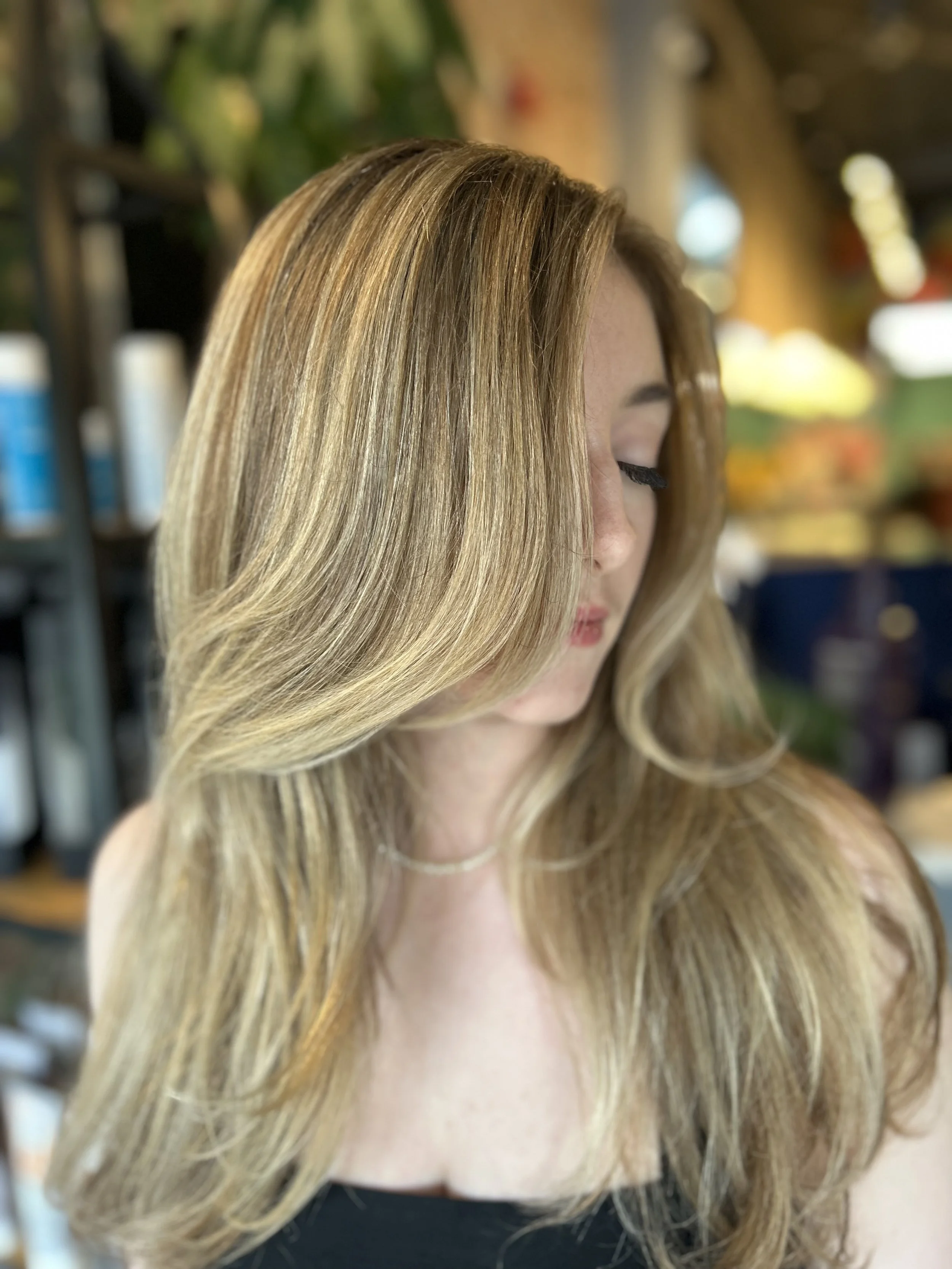 A woman with lightly curled, blonde hair, looking down with her face partially obscured by her hair, in a salon or similar setting with blurred shelves and bottles in the background.