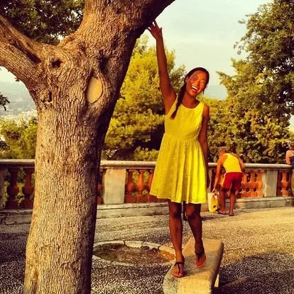 A woman in a yellow dress smiling and posing with one arm raised, leaning against a large tree on a sunny day, with a stone railing and other people in the background.