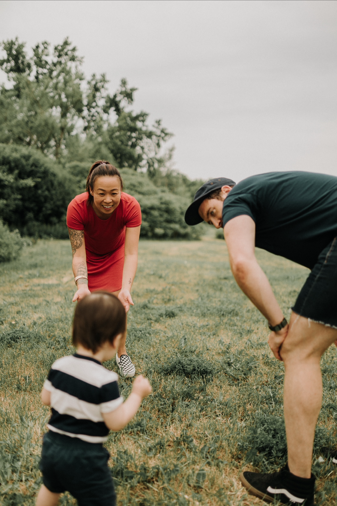 A woman and a man are playing with a young child outdoors on a grassy field, all smiling and bending forward.