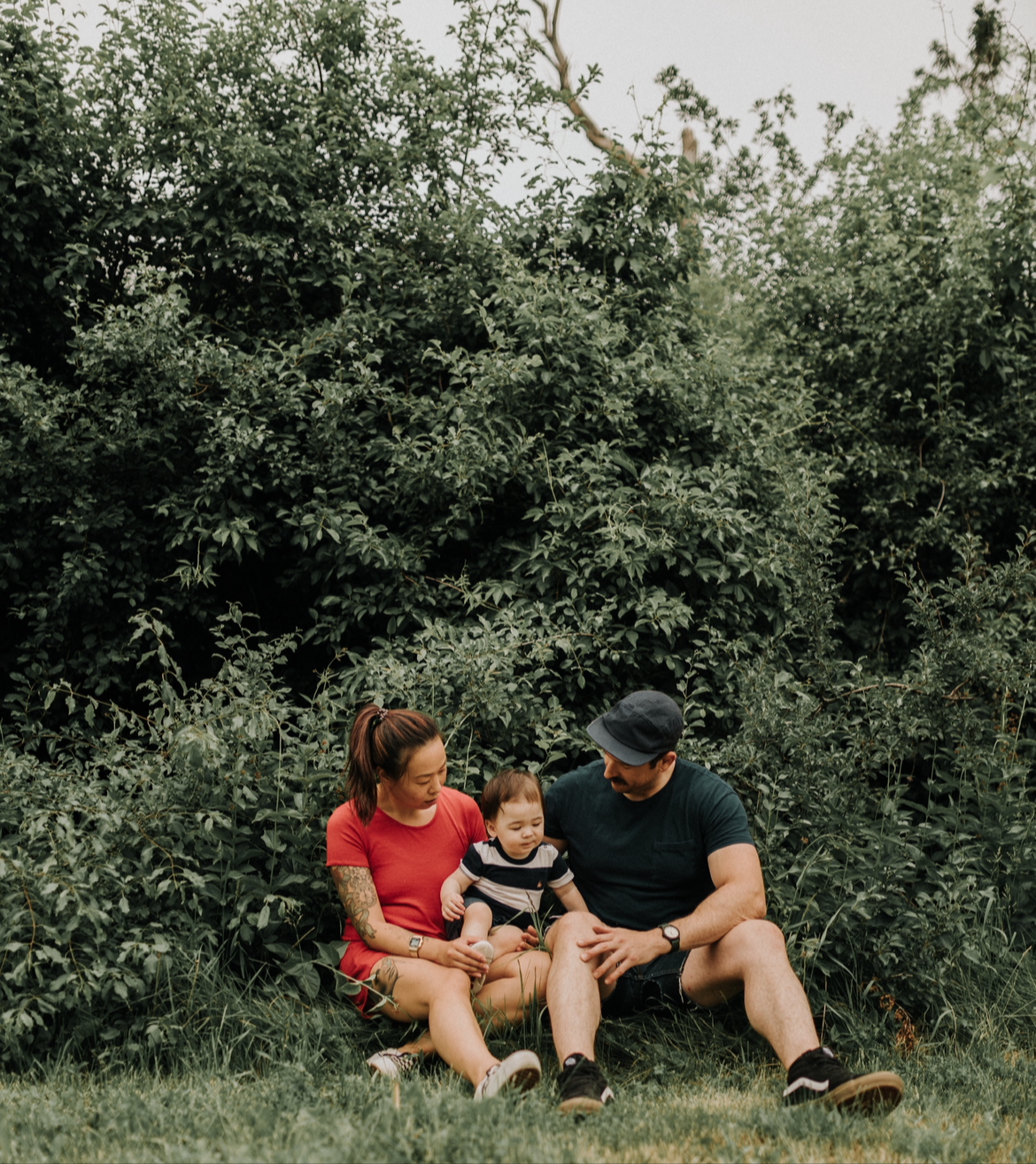 A family of three sitting on the grass in front of dense green bushes, with a woman in a red dress, a man in a black shirt and hat, and a young child in a striped shirt.