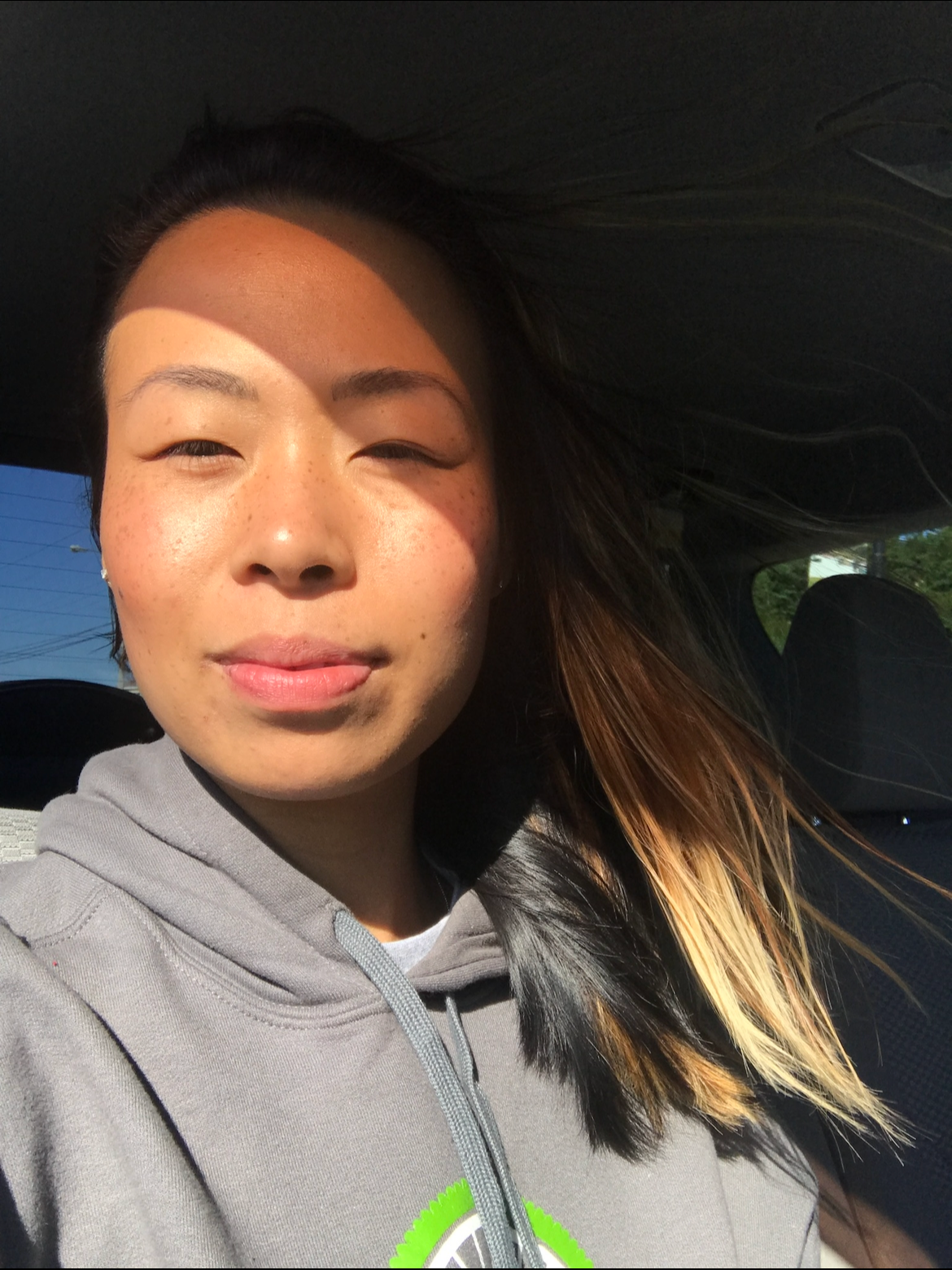 A woman with long dark hair and freckles taking a selfie inside a car on a sunny day.