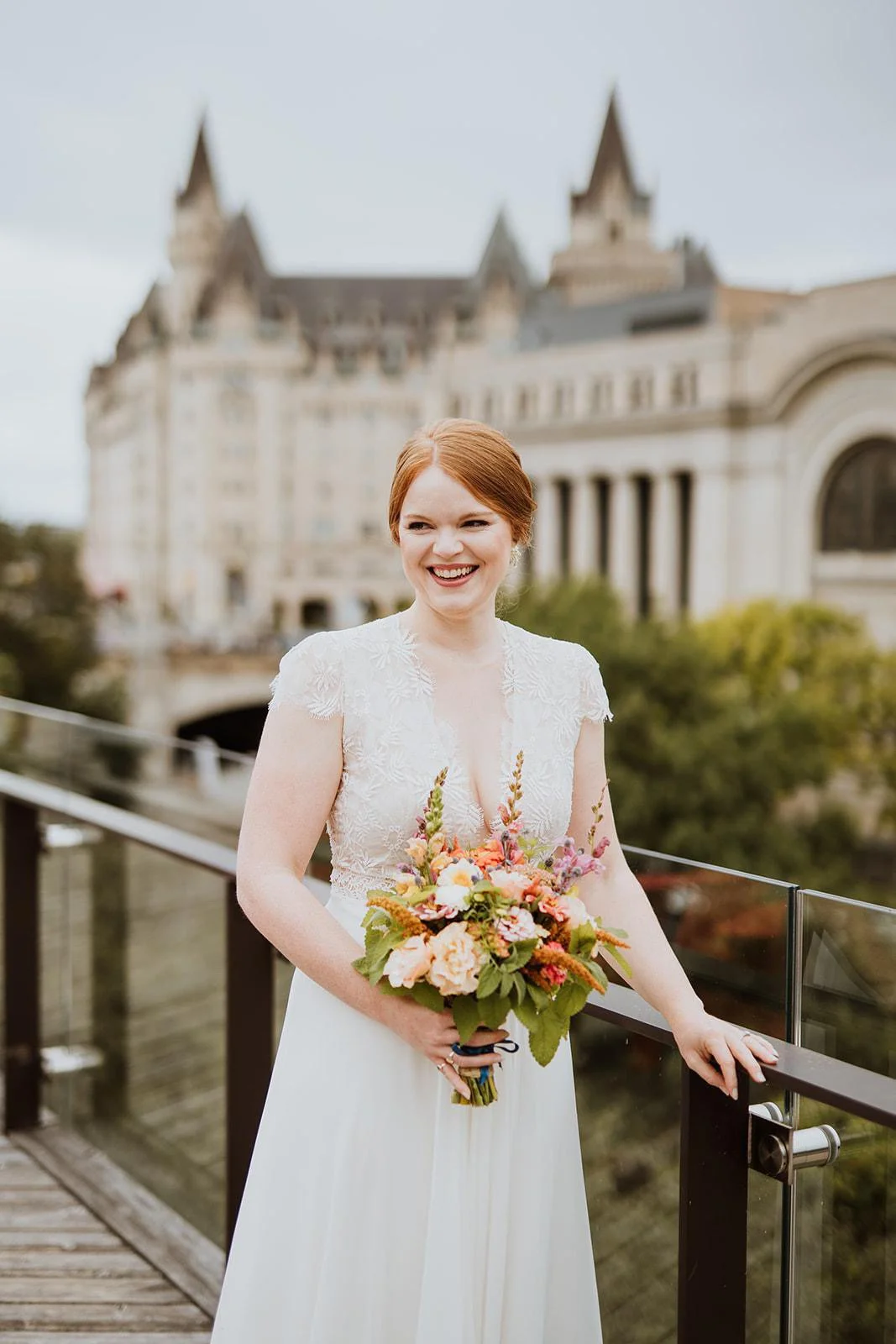 A smiling bride with red hair in a white dress holding a bouquet of flowers, standing on a balcony with a castle-like building in the background.