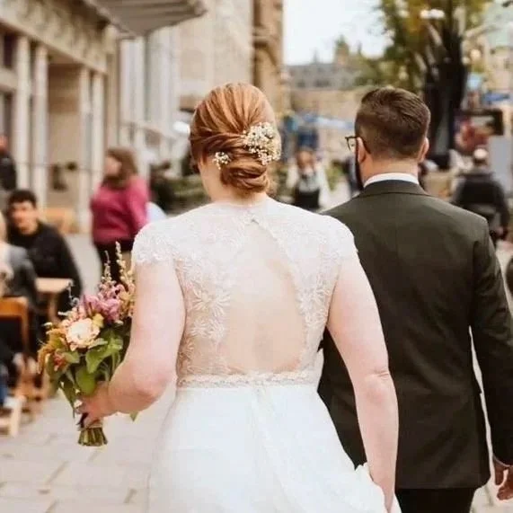 Back view of a bride holding a bouquet walking with her groom on a city street, with pedestrians and buildings in the background.